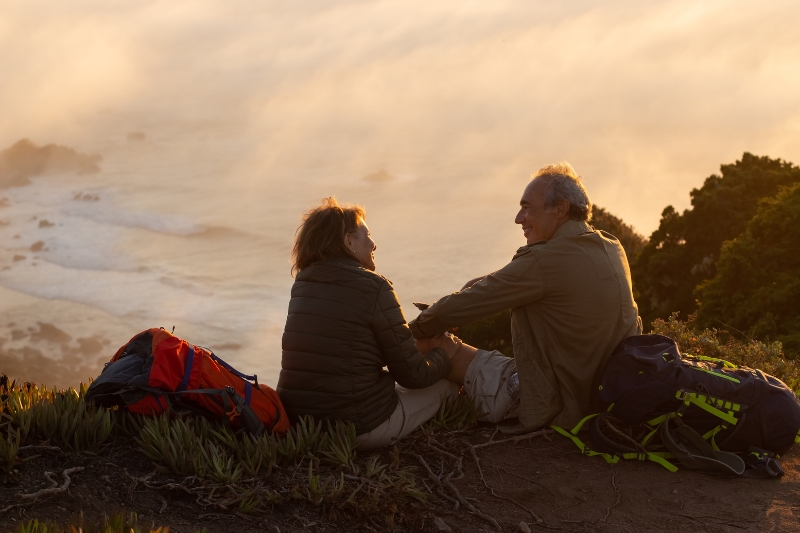Una pareja senior practica turismo slow y disfrutan de las vistas desde un mirador cerca del mar y la playa al atardecer. 